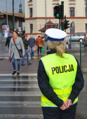 Policjantka stoi przed przejściem dla pieszych z sygnalizacją świetlną. Przez przejście przechodzą piesi.