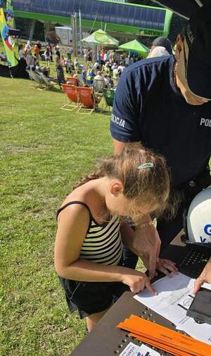 Umundurowany policjant na stoisku profilaktycznym i dziewczynka odbijająca linie papilarne na pamiątkowym dyplomie