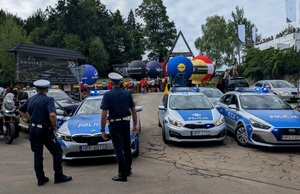 Na zdjęciu trzy radiowozy oraz dwóch policjantów stojących tyłem. W tle kibice