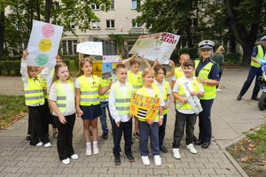Policjantka oraz dzieci ubrane w kamizelki odblaskowe z transparentami z hasłami bezpieczeństwa na drodze