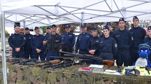 Kierownictwo jednostki oraz umundurowani policjanci i ucznioiwe klasy o profilu policyjnym stojący przy stole z bronią kolekcjonerską oraz ulotkami informacyjnymi