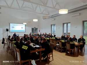 strażacy siedzą przy stołach na zajęciach z policjantem