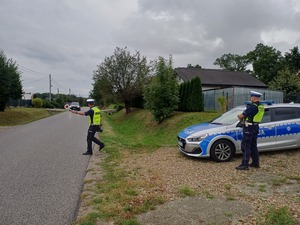 Dwóch policjantów przy radiowozie na poboczu drogi. Jeden policjant zatrzymuje jadący pojazd.