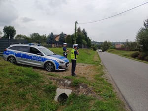 Dwóch policjantów przy radiowozie na poboczu drogi. jeden polcjant mierzy prędkość jadącym samochodom.