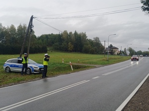 Dwóch policjantów stoi na poboczu drogi obok radiowozu. Jeden z nich mierzy radarem prędkość jadących pojazdów.