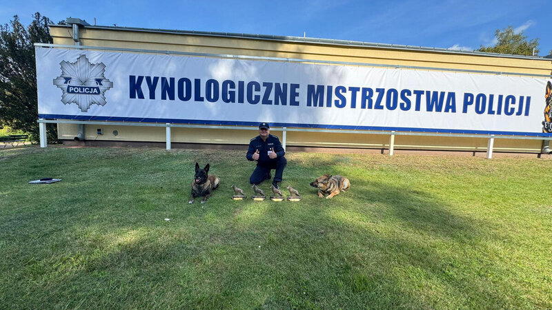 policjant z psami służbowymi oraz pamiatkowymi statuetkami na tle baneru Kyneologiczne mistrzostwa policji