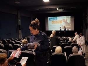 policjantka rozdaje ulotki seniorom siedzącym na fotelach przed prelekcją
