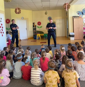 Dwóch umundurownych policjantów, przed nimi dzieci siedzące na dywanie