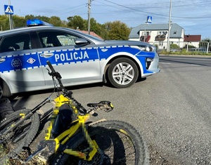 Żółty rower położony na poboczu drogi, w tle radiowóz