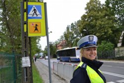 Zdjęcie przedstawia policjantkę ruchu drogowego na tle znaków: D-6 – przejście dla pieszych oraz T-27 tzw. Agatka – tabliczka wskazująca, że przejście dla pieszych jest szczególnie uczęszczane przez dzieci.