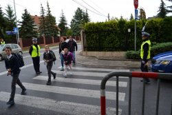 Zdjęcie przedstawia skrzyżowanie z przejściem dla pieszych. Po dwóch stronach przejścia dla pieszych policjantki ruchu drogowego wstrzymują ruch pojazdów – jedna policjantka trzyma w ręku podniesioną do góry tarczę do zatrzymywania pojazdów. Przez przejście dla pieszych przechodzą dzieci wraz z rodzicami (2 chłopców i 1 dziewczynka).