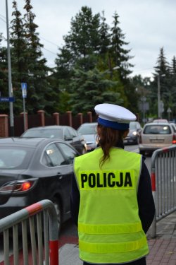Zdjęcie przedstawia policjantkę  ruchu drogowego stojącą tyłem do obiektywu a przodem w kierunku drogi Na plecach widoczny duży napis „Policja”. W tle – wzmożony ruch drogowy.