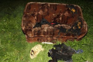 kanapa koloru brązowego ze śladami podpalenia leżąca na trawniku, przed kanapą pozostałości spalonych ubrań