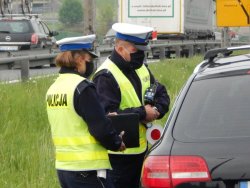 Zdjęcie przedstawia policjantkę i policjanta ruchu drogowego podczas kontroli kierującego pojazdem. Policjant trzyma w ręku ręczny miernik prędkości, a policjanta trzyma w ręku notes. W tle widoczne są samochody jadące jezdną, która rozdzielona jest pasem zieleni.