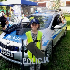 dziewczynka stoi przed radiowozem, ma na sobie elementy umundurowania i wyposażenia policjanta