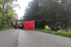 Na zdjęciu widać masyw leśny, a w nim wywrócony pojazd. Na drodze stoi wóz strażacki i rozłożony czerwony parawan. Na miejscu zdarzenia umundurowani policjanci ruchu drogowego wykonują czynności.