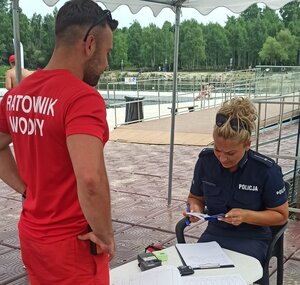 Policjantka umundurowana dokonuje kontroli książeczki uprawnień ratowniczych, ratownikowi stojącemu nad policjantka ubrany w czerwony t-shirt z napisem ratownik wodny