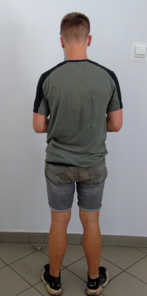 Zatrzymany nieletni, stojący tyłem. Włosy krótkie ciemny blond, ubrany w koszulkę koloru zielonego z czarnym pasem na ramionach i ciemne dżinsowe krótkie spodenki.