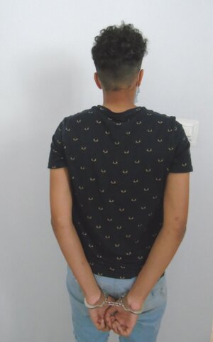 Zatrzymany nieletni, stojący tyłem, na rękach trzymanych z tyłu ma załozone kajdanki. Włosy krótkie ciemne, ubrany w czarną koszulkę i niebieskie dżinsy