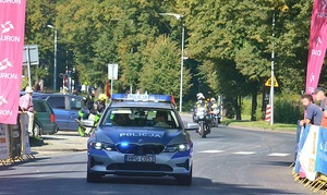Oznakowany radiowóz jedzie wzdłuż trasy kolarzy a obok metalowe barierki i stojącymi za nimi kibicami oczekującymi na peleton