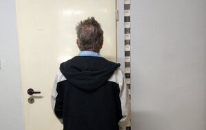 Zatrzymany mężczyzna ubrany w czarno-białą kurtkę z kapturem oraz niebieską koszule stojący przed wskaźnikiem wzrostu oraz białymi drzwiami