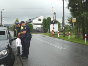 Umundurowany policjant ruchu drogowego oraz pracownik Polskich Linii Kolejowych stojący obok samochodu, trzymający w ręku ulotki