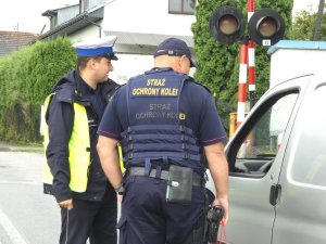Umundurowany policjant ruchu drogowego oraz Straży Ochrony Kolei, którzy stoją obok samochodu w rejonie przejazdu kolejowego
