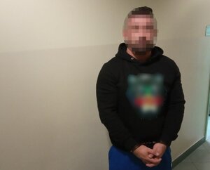 Zatrzymany mężczyzna ubrany w czarną bluzę z kapturem z kolorowymi elementami z rękami zakutymi w kajdanki stojący przodem do zdjęcia