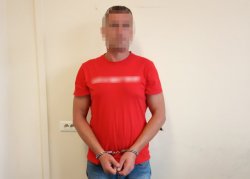 Zatrzymany mężczyzna ubrany w czerwoną koszulkę z rękami zakutymi w kajdanki stojący przodem do zdjęcia