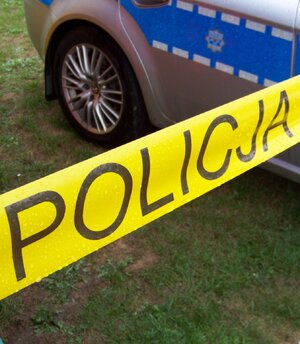 fragment żółtej taśmy z napisem Policja a w tle oznakowany radiowóz