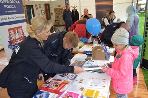 Policjantka wręcza dziewczynce element odblaskowy. Policjant przybija pieczątkę na kartkę. W tle inne osoby znajdujące się przy policyjnym stanowisku.