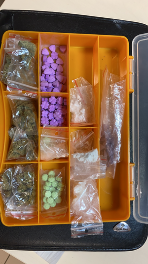 Kilka woreczków foliowych z zawartością suszu roślinnego, białego proszku oraz folią aluminiową. Tabletki w kolorze zielonym i fioletowym. Rzeczy znajdują się w żółtym plastikowym pojemniku.