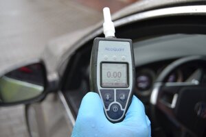 W ręku trzymany jest alkomat , na którym wyświetlony jest wynik 0.00. W tle widoczny fragment wnętrza pojazdu.