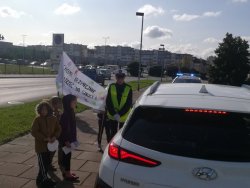 Na zdjęciu policjantka ruchu drogowego wraz z uczniami rozmawia z kontrolowanym znajdującym się w pojeździe. Dzieci mają e sobą transparent z napisem „Chcemy bezpiecznie dotrzeć do szkoły”.