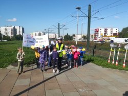 Na zdjęciu grupka dzieci wraz z policjantką ruchu drogowego stojąc na chodniku promują bezpieczeństwo na drodze, trzymając w ręku transparent z napisem „Chcemy bezpiecznie dotrzeć do szkoły”.