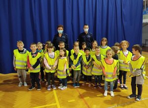 Grupa dzieci w kamizelkach odblaskowych, które stoją przodem. Za nimi umundurowana policjantka i policjant.