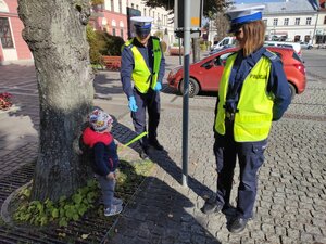 Policjant i policjantka ruchu drogowego w odblaskowych kamizelkach. Policjant wręcza odblask dziecku. W tle widać olkuski Rynek, sklepy i budynki oraz zaparkowane samochody. Pogoda słoneczna.