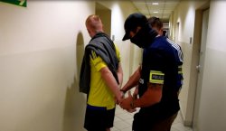 policjanci kryminalni zakładają kajdanki na ręce trzymane z tyłu zatrzymanemu ubranemu w żółtą koszulkę z zarzucona na ramię szarą bluzą