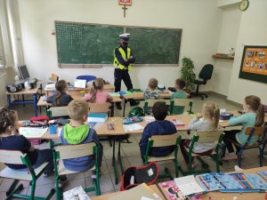 Policjant ruchu drogowego w białej czapce i odblaskowej kamizelce prowadzi zajęcia w klasie z uczniami. Dzieci siedzą w szkolnych ławkach.