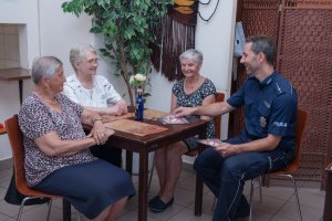 Trzy kobiety siedzą przy stoliku, a Policjant wręcza im ulotki  dotyczące bezpieczeństwa seniorów.