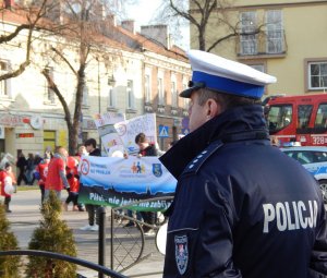 Policjant ruchu drogowego (biała czapka, widoczny napis POLICJA na plecach kurtki) w stopniu nadkomisarza, w tle młodzież z transparentami nawołującymi do jazdy w trzeźwości, przygotowująca się do przemarszu