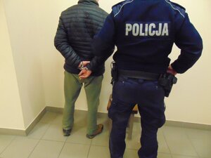 mężczyzna z założonymi na ręce trzymane z tyłu kajdankami, a przy nim policjant
