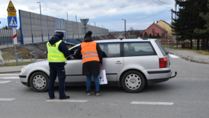 Policjant w kamizelce odblaskowej z napisem POLICJA oraz pracownik PKP Polskich Linii Kolejowych wręczają ulotki przy pojeździe osobowym