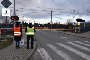 Policjant w kamizelce odblaskowej z napisem POLICJA oraz pracownik PKP Polskich Linii Kolejowych stoją przed zamkniętym przez rogatki przejazdem kolejowo-drogowym
