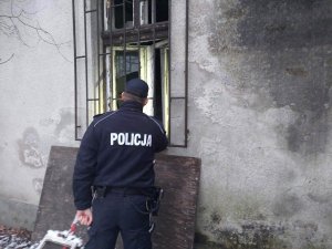Umundurowany policjant stojący tyłem zerkający przez okratowane okno do wnętrza budynku