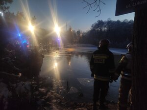 Staw w Wolbromiu. Dookoła stawu widać wozy strażackie i policyjne na włączonych sygnałach oraz pracujących przy akcji strażaków i policjantów. Zdjęcie wykonane nocą, porą zimową. Na wodzie widać gdzieniegdzie warstwę cienkiego lodu