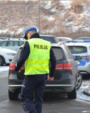 Policjant ruchu drogowego (widoczna biała czapka, kamizelka odblaskowa z napisem na plecach POLICJA) podchodzący do zatrzymanego do kontroli samochodu. W tle widać zimową aurę.