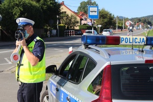 Umundurowany policjant z ruchu drogowego mierzący prędkość przy pomocy miernika stojący przy drodze wojewódzkiej po której poruszają się samochody