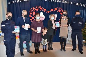 dziadkowie z wnuczką, policjanci, pani dyrektor przedszkola i pani z zarządu województwa trzymają w rękach kartki dla seniorów przestrzegające ich przed oszustami