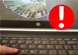 cyberoszustwa - klawiatura laptopa, na ekranie widoczne pieniądze, obok czerwony znak z wykrzyknikiem